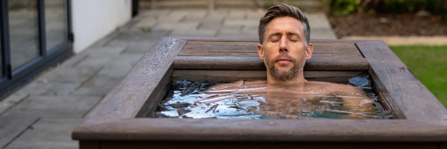 Man relaxing in a wooden ice bath tub outdoors during a cold plunge for recovery and wellness benefits.