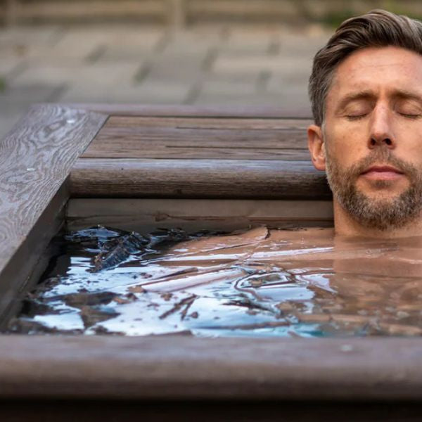 Man relaxing in a wooden ice bath tub outdoors during a cold plunge for recovery and wellness benefits.