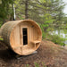 Wooden barrel-shaped sauna in a forest setting with trees and water in the background