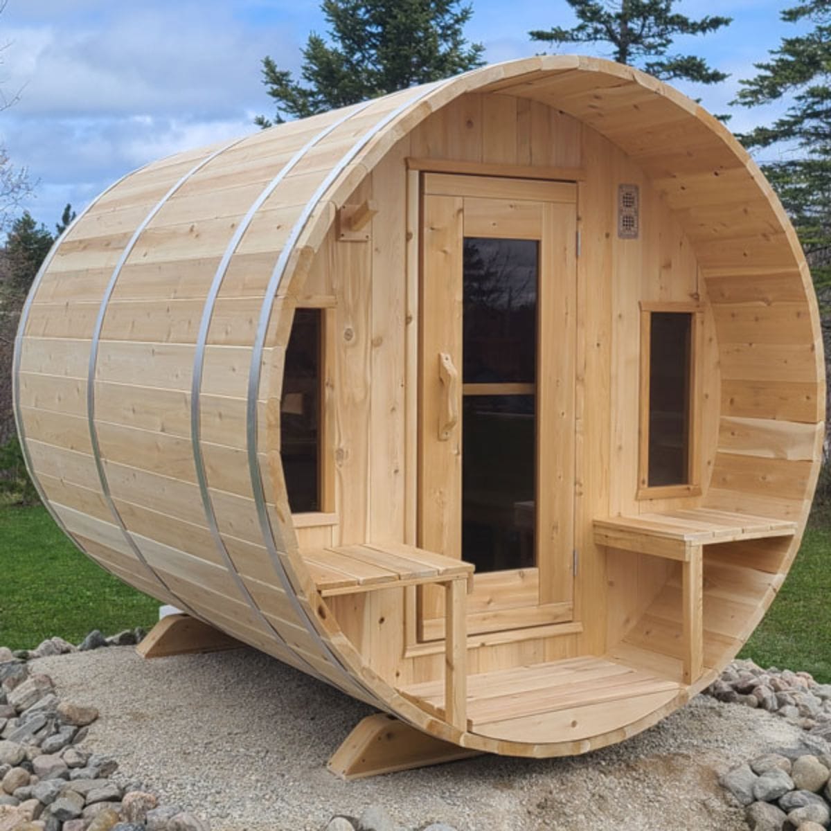 Wooden barrel-shaped sauna with a door and windows, set outdoors on a gravel surface with trees in the background.
