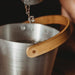 A stainless bucket with a wooden handle is being filled with liquid from a metal pitcher