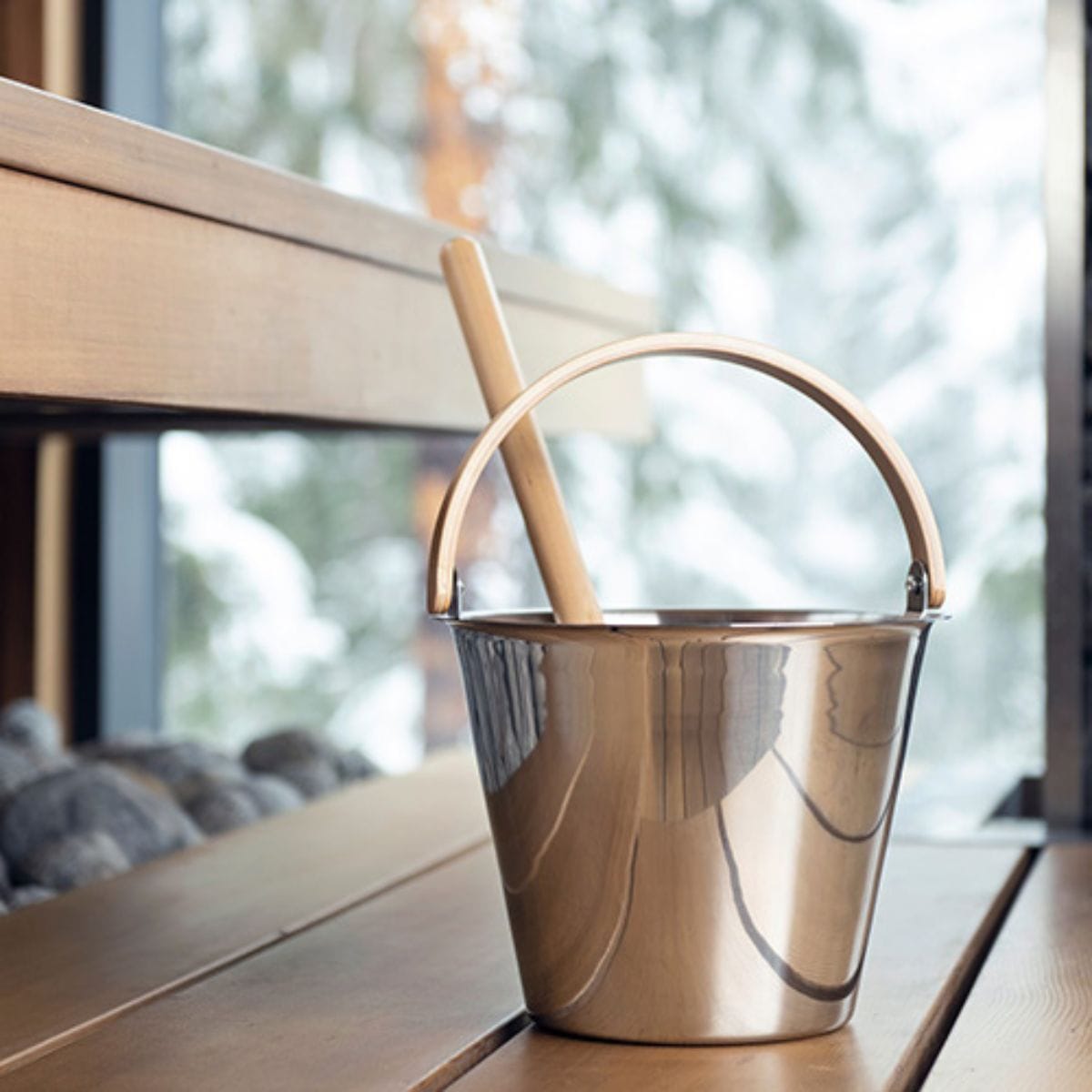 Metal bucket with a wooden handle on a wooden surface near a window with a blurred greenery background