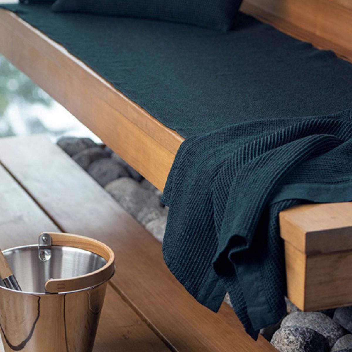 Wooden bench with dark green cushion and blanket, next to a metal cup on a wooden surface.