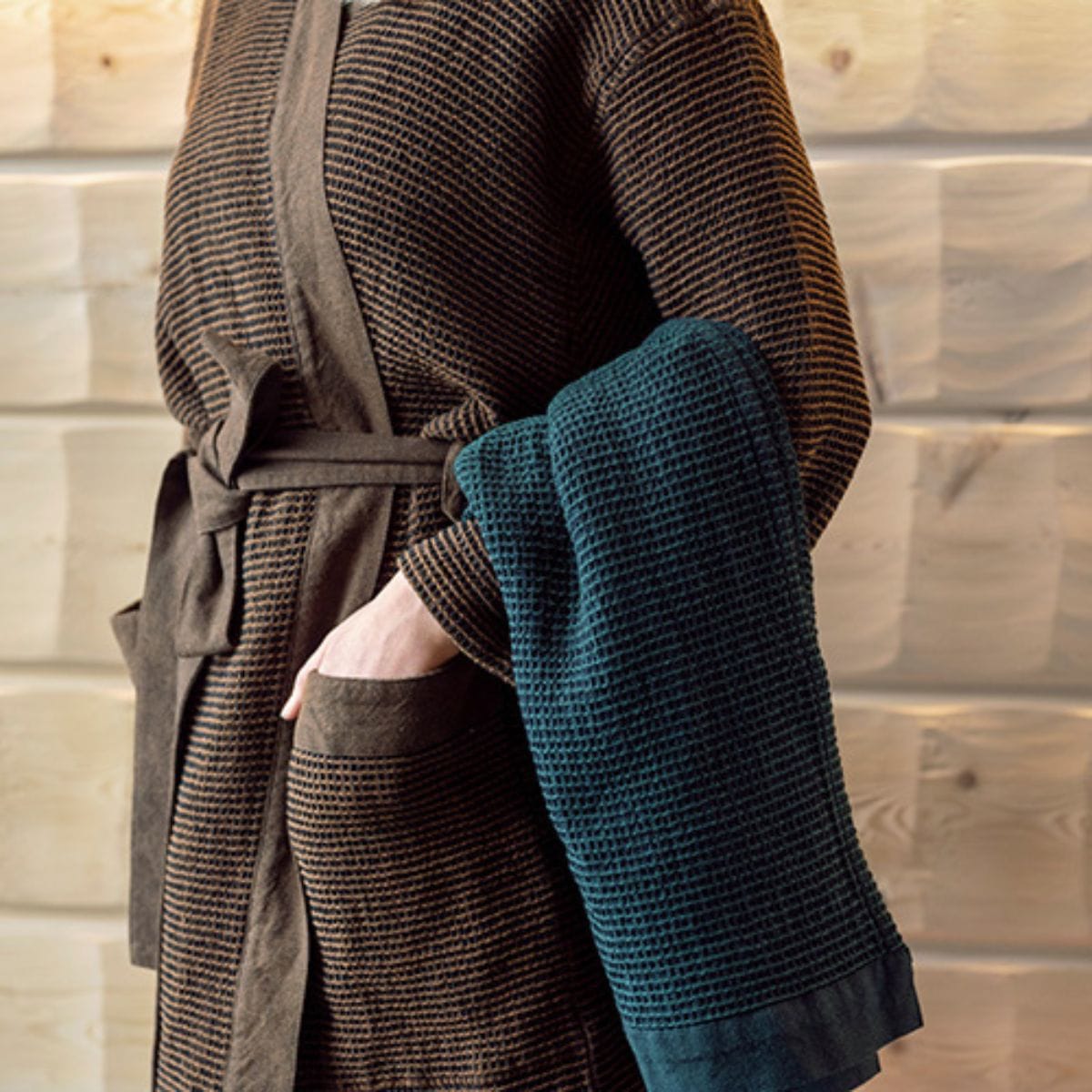 Person wearing a textured robe holding a matching towel against a wooden wall.